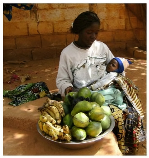 une jeune femme portant un bébé, devant un grand plat de fruits (papayes, bananes)