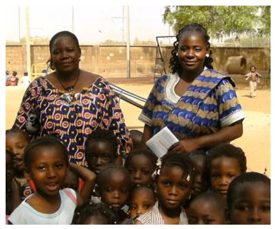 deux femmes (une enseignante et la Directrice Mamounata) entourées d'enfants dans la cour de la maternelle du Camp